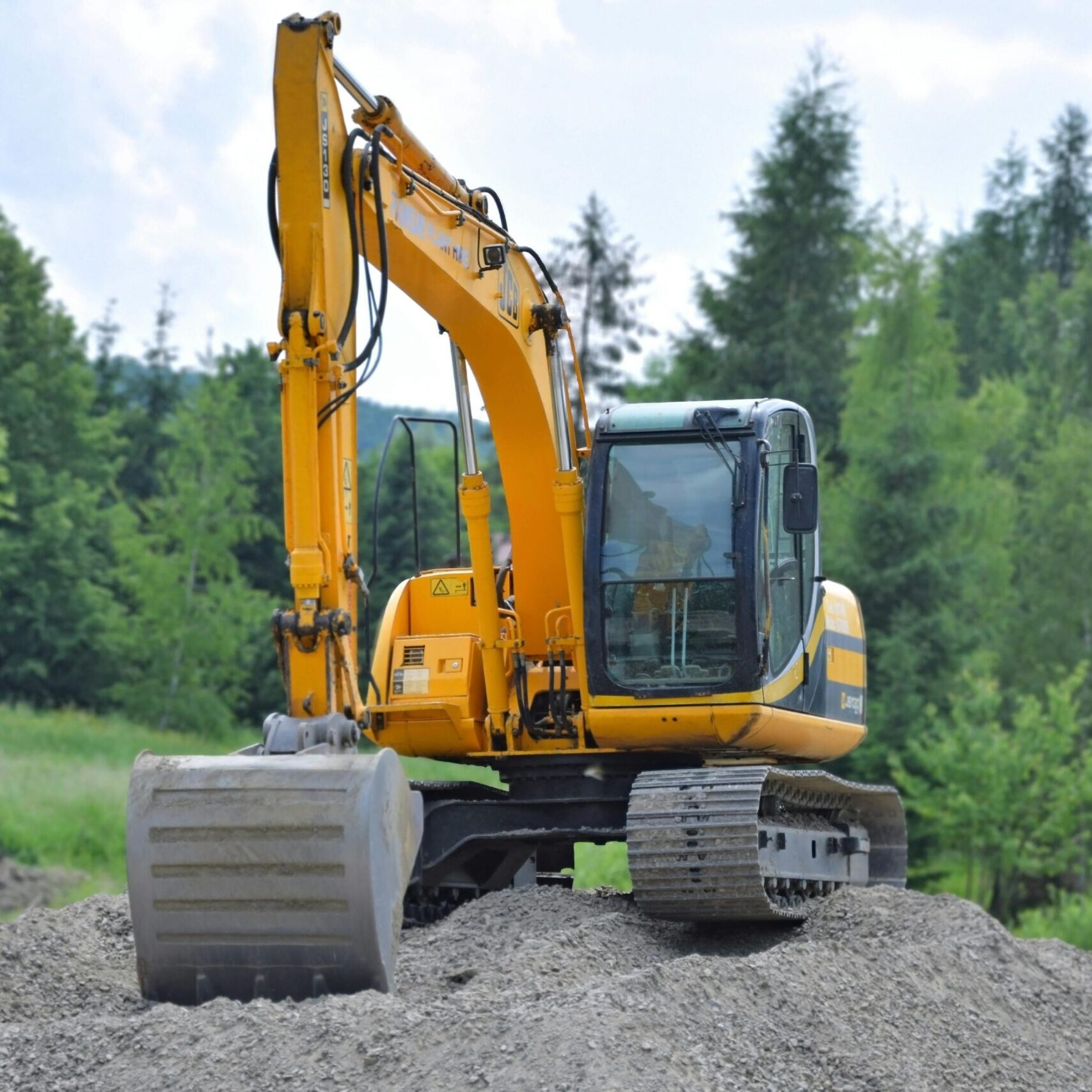 Inicio A yellow excavator on a pile of gravel in a lush, green forest setting in Poland.